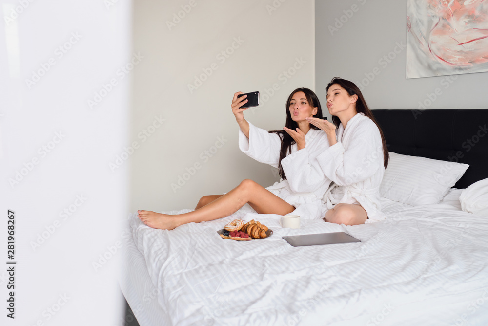 Wall mural Two attractive women in white bathrobes sending air kisses dreamily taking photos together on cellphone. Beautiful girls having breakfast in bed with laptop in hotel