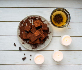 handmade souffle cubes covered with cocoa powder lie on a plate on a light wooden table, hot tea in a glass mug and next to it three white burning candles, pieces of chocolate and a sprig.