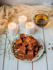 handmade souffle cubes covered with cocoa powder lie on a plate on a light wooden table, hot tea in a glass mug and next to it three white burning candles, pieces of chocolate