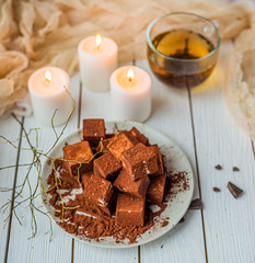 handmade souffle cubes covered with cocoa powder lie on a plate on a light wooden table, hot tea in a glass mug and next to it three white burning candles, pieces of chocolate