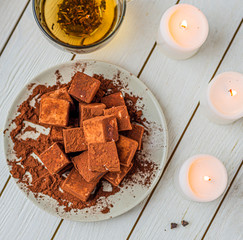handmade souffle cubes covered with cocoa powder lie on a plate on a light wooden table, hot tea in a glass mug and next to it three white burning candles, pieces of chocolate