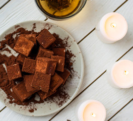 handmade souffle cubes covered with cocoa powder lie on a plate on a light wooden table, hot tea in a glass mug and next to it three white burning candles, pieces of chocolate