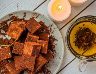 handmade souffle cubes covered with cocoa powder lie on a plate on a light wooden table, hot tea in a glass mug and next to it three white burning candles, pieces of chocolate
