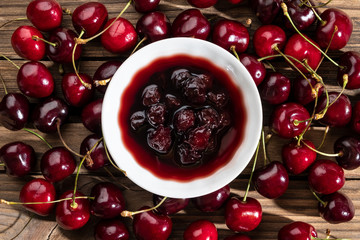 Cherry jam in a small bowl close-up on a wooden background, spread around a lot of ripe juicy cherries