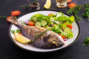 Baked fish dorado with lemon and fresh salad in white plate on dark rustic background. Healthy dinner with fish concept. Dieting and clean eating