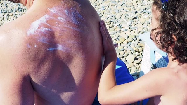 4K Little son applying sunscreen lotion on dads back at beach. Family preparing for bath in sea. Caring and safety concept. Slow Motion