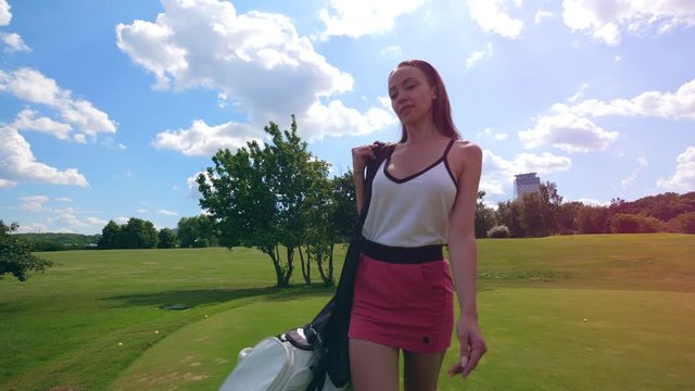 Young woman walks on a golf course, holding a bag with equipment.