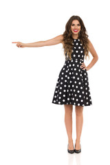 Beautiful Young Woman In Black Cocktail Dress In Polka Dots And High Heels Is Pointing And Smiling
