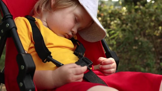 Close up outdoor portrait of little blonde boy in cap sleeping sweet in red carriage in summer beautiful forest with sun shining on background. Child health care concept. Traveling lifestyle.