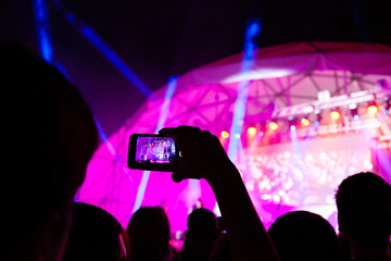 Fototapeta na wymiar Raised hands of a man with a cell phone with a digital screen in front of bright lights record a concert. Unrecognizable people take photos and videos on the phone in front of the stage. Night show.