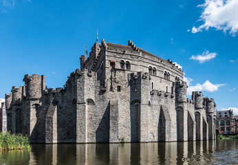 Fototapeta na wymiar Gent, Flanders, Belgium - June 21, 2019: Gray stone castle and ramparts of Gravensteen, historic medieval castle of city, behind its moad against blue sky with white clouds. Flags on top.