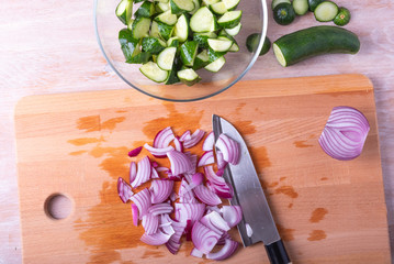 Sliced red onions and santoku knife on a cutting board and sliced cucumbers in a bowl for salad.