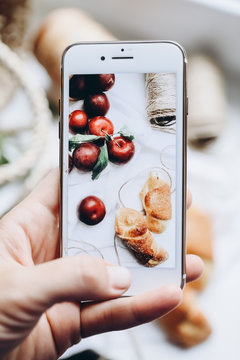 A hand holds a smartphone and takes a still life picture of juicy plums and croissants