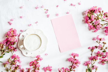 Morning coffee mug for breakfast, empty notebook and pink flowers on white wooden table, top view, flat lay style. Woman working desk. Mockup, overhead