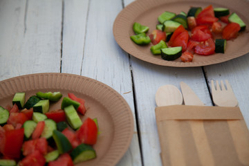 paper cardboard dishes with vegetable salad tomatoes cucumbers with wooden devices fork horse knife on a white wooden table