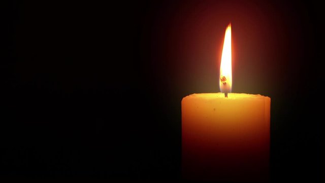 Illuminated Candle Loop is a stock video with a lit flame that flickers and glows in a dark environment. The wax is white or light yellow.