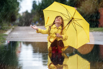 Happy funny child girl with umbrella jumping on puddles in rubber boots