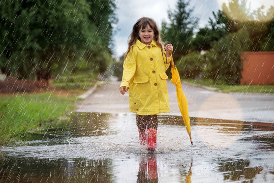 Happy funny child girl with umbrella jumping on puddles in rubber boots