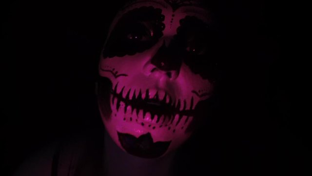 Halloween mask CALAVERA CATRINA. A girl in a blinking and frightening light. Mexican day of the dead. Portrait of a young woman with scary multi-colored makeup for Halloween on a dark background. 4K