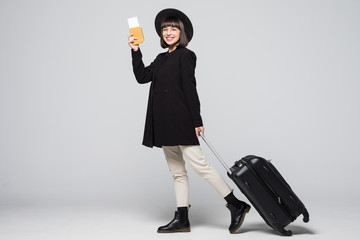Happy young woman in coat and hat walking with the travel bag, holding passport and tickets isolated on white background