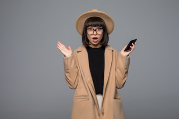 Smiling woman shocked use smartphone in beige coat, round hat on gray background