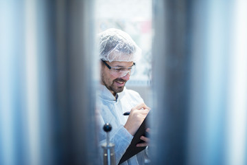 Technologist in white protective suit standing by industrial machines and writing report on quality...