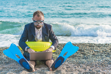 Successful businessman wearing snorkeling tools is working on the beach with laptop..