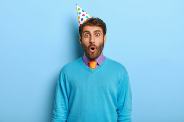 Portrait of shocked unshaven guy wears party hat, blue sweater with purple shirt and tie, surprised...