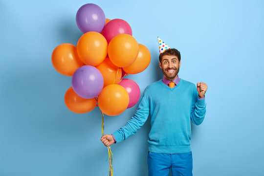 Birthday party concept. Positive man clenches fists with happiness, holds bunch of multicolored balloons, wears holiday cap and blue festive clothes, stands indoor, waits for guests or friends