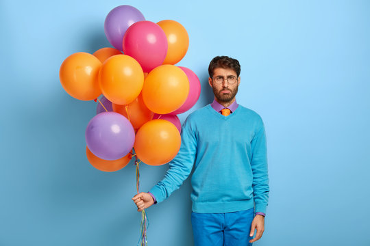 Angry Discontent Man Has No Wish To Go On Party, Grieved Facial Expression, Holds Colorful Balloons In Hand, Dressed In Fashionable Apparel, Isolated On Blue Background. Lonely Celebration Concept