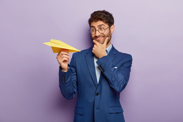 Prosperous young company owner has goal, touches chin and looks happily at hand made paper plane,...