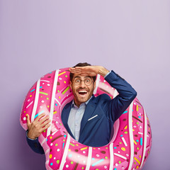 Vertical image of happy male entrepreneur gazes into distance, keeps palm near forehead, wears...