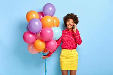 Fototapeta na wymiar Satisfied beautiful Afro American birthday girl has pleasant conversation, receives congratulation from relatives and friends, wears red shirt, yellow skirt, holds party attributes, celebrates holiday