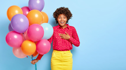Fototapeta na wymiar Half length shot of cheerful surprised woman touches chest, enjoys balloons theme party, has fun, reacts on awesome present, wears stylish red shirt and yellow skirt, poses indoor over blue wall