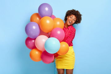 Fototapeta na wymiar Happiness and celebration concept. Overjoyed dark skinned female greets friends with special occasion, holds huge bunch of helium balloons, has cheerful moments in her life during birthday party