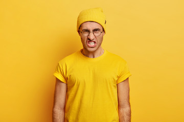 Portrait of irritated man looses temper, clenches teeth from anger, wrinkles nose, looks with hate at camera, says you piss me off, wears transparent glasses, yellow clothes, stands indoor, argues