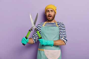 Dissatisfied landscpae designer or gardener holds gardening shears, going to cut green bush, wears...