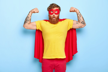 Emotive surprised male hero has noble qualities, demonstrates strength with raised arms, has strong muscles, dressed in superhero costume, isolated on blue, has strength to face dire situation in life - obrazy, fototapety, plakaty