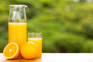 Orange juice in a jug and a glass with full and cut oranges by a window opening to tree tops, shallow depth of field. large copy space.