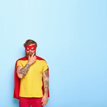 Vertical image of thoughtful superhero thinks how to do something nice for someone, wants to make right thing, focused away, wears red mask and cloak, has tattooed arms, isolated on blue wall