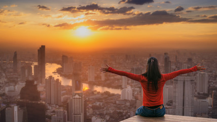 Fototapeta na wymiar Rear view of female traveler with raised hands standing on the top of a tower and looking at the urban city with sunset sky. Woman happy enjoying the city view from top of a city at evening.