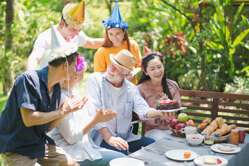 Happy big family have a lunch at outdoor in green garden. Grand father's birthday party on picnic table in summer. Old man hold the cake. Big family outdoor lunch party concept.