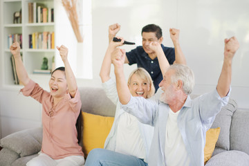 Group of positive seniors European and Asia are sitting on the big sofa and talking which looking television at retirement home. Retired old people are smiling and felling happy.