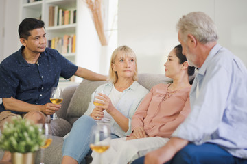 Obraz premium Group of positive seniors European and Asia are talking and drinking wine which sitting on sofa in living room at retirement home. Retired people have felling be serious.