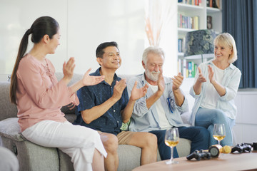 Obraz premium Group of positive seniors European and Asia are singing and drinking wine which sitting on sofa in living room at retirement home. Retired people have smiling and felling happy.