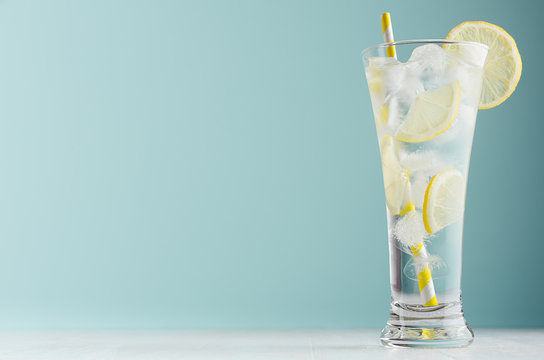 Detox cold tonic water with sunny lemon slices, ice cubes, yellow striped straw in elegant glass on white wood table, mint color wall.