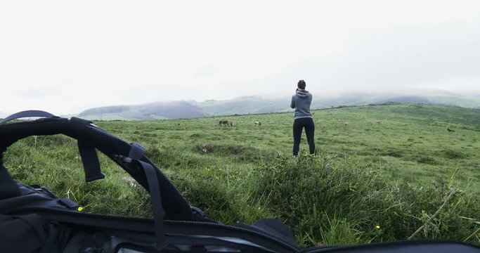 Adventurous girl taking photos of horses on a foggy spring day in the mountains. Backpack in the foreground. Handheld stabilized shot.