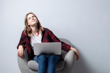 young girl with a laptop sitting on a soft comfortable chair and thinking, a woman using a computer against a white blank wall, she freelancing and dreaming, copy space