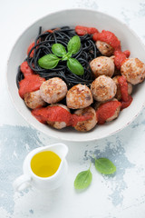 Bowl of squid-ink spaghetti with pork meatballs, tomato sauce and fresh green basil, elevated view on a white concrete surface
