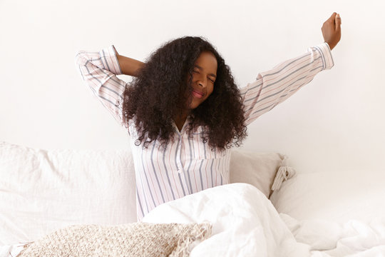 Sleepy pretty girl with brown skin and voluminous hairdo stretching arms, keeping eyes closed, feeling lazy to get up early in the morning. Portrait of stylish cute young African woman in bed
